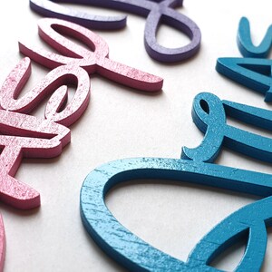 May include: Three wooden names, "Casey", "Elle", and "Ava", are painted in pink, purple, and teal.