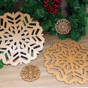 May include: Three wooden snowflake coasters in different sizes. The coasters are made of light brown wood and have a delicate snowflake design cut out of them. The coasters are arranged on a wooden surface with a green garland in the background.