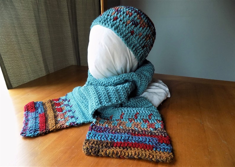 May include: A teal blue crocheted hat and matching scarf with brown, red, and purple accents. The hat and scarf are made with a textured stitch.