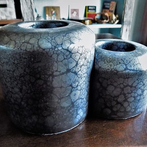 May include: Two black ceramic candle holders with a crackled glaze finish. The holders are cylindrical in shape and have a slightly rounded top.