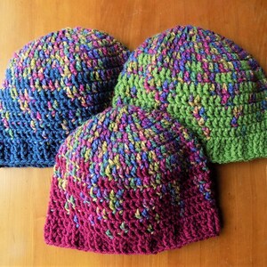 May include: Three crocheted hats in different colors. The hats are made with a multi-colored yarn with shades of blue, green, pink, and yellow. The hats are stacked on top of each other.