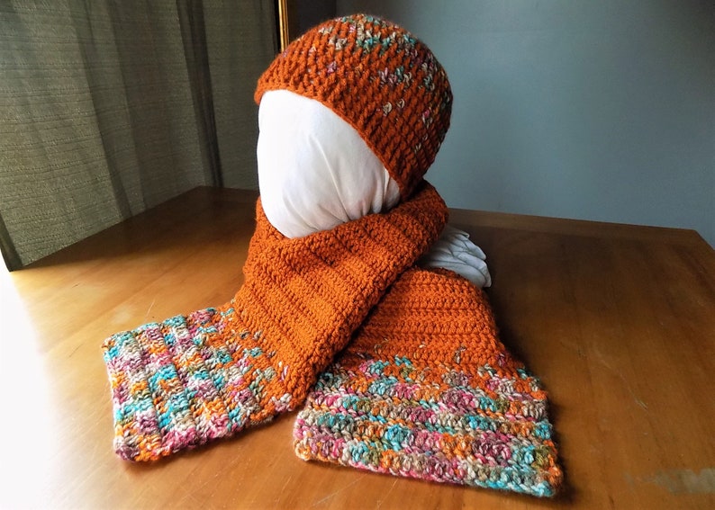May include: A crocheted hat and scarf set in a rust orange color with a multi-colored flecked pattern. The hat is a slouchy beanie style and the scarf is long and rectangular.