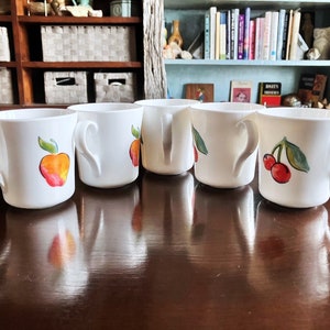May include: Five white ceramic mugs with painted fruit designs. The mugs have a rounded shape and a handle on the side. The designs include a red apple, a yellow pear, a red cherry, and a green leaf.