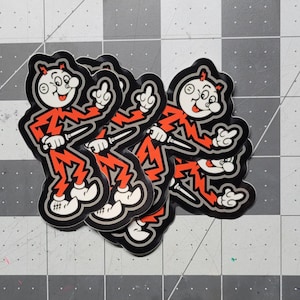Op de afbeelding: Een stapel van vijf zwarte en rode stickers met een cartoonfiguur met een bliksemontwerp. Het personage steekt de middelvinger op en houdt een mes vast.