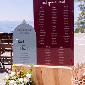 May include: A burgundy rectangular acrylic seating chart with white text titled find your seat and a light gray arched acrylic welcome sign with the text welcome to our love story, Karl and Charlotte, and the date 09.20.2026 on a wooden base.