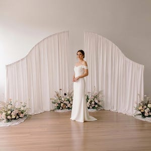 May include: A floor-length white off-the-shoulder gown with a fitted bodice and a subtle train. The garment is positioned in front of two large, arched white fabric backdrops with gathered drapes, flanked by three floral arrangements.
