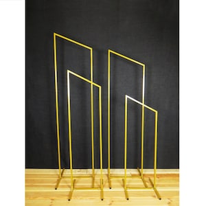 May include: Three gold metal archways of different sizes, standing on a wooden floor. The archways are made of thin metal bars and are designed to be used as a backdrop for events or photos.