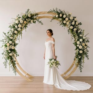May include: A large circular wooden arch decorated with white roses, cream-coloured flowers, and green eucalyptus foliage. The arch stands on a light-coloured floor and frames a floor-length white gown with off-the-shoulder sleeves and a train.