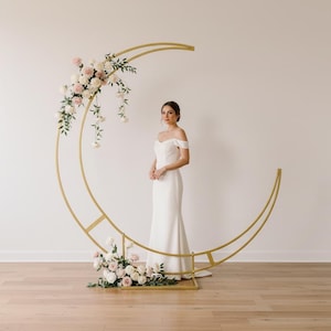 Könnte beinhalten: Ein goldfarbener, mondförmiger Hochzeitsbogen, verziert mit weißen und rosa Blumen. Eine Person in einem weißen schulterfreien Kleid steht vor dem Bogen. Der Bogen steht auf einem Holzboden.