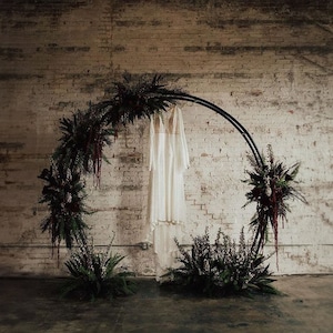 May include: A white wedding dress hangs on a black metal hoop decorated with dark green and burgundy floral arrangements against a white brick wall.