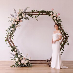 May include: A large hexagonal wooden wedding arch decorated with white and pale pink roses, blue delphinium, and green ivy vines. A person stands to the right of the arch wearing a floor-length white off-the-shoulder gown with a fitted bodice.