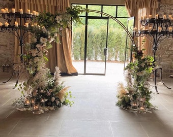 May include: An elegant wedding arch decorated with white flowers, pampas grass, and greenery. The arch is made of gold metal and is set in front of a glass door. Candle holders with lit candles flank the arch.