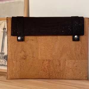 Può includere: Una custodia per laptop in sughero e pelle marrone con chiusura a lembo e due bottoni a pressione in metallo. La custodia è realizzata in sughero naturale e ha un lembo in pelle marrone con un cinturino in pelle marrone.