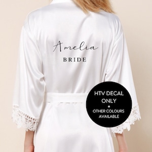 May include: White satin robe with lace trim on the sleeves. The back of the robe has the text "Amelia Bride" printed in black.