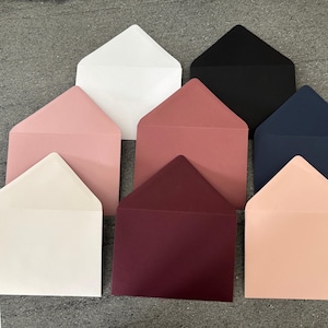 May include: A selection of coloured envelopes in shades of white, pink, burgundy, black, and navy blue. The envelopes are arranged on a grey surface, displaying their different hues and textures. The envelopes are blank and ready for use.