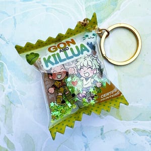 May include: A clear acrylic snack bag keychain featuring a gold metal ring. The bag has green serrated edges, green star-shaped glitter inside, and a graphic of two cartoon characters with the text GON AND KILLUA and CERTIFIED HUNTERS.