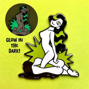Pode incluir: Um broche de esmalte preto e branco de uma mulher com cabelo verde sentada em uma explosão verde e preta. O broche brilha no escuro. "Glow in the dark!" está escrito abaixo do broche.