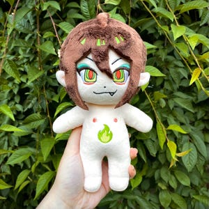May include: A plush doll with short brown hair, bright green eyes, and a small fang. The doll has a white body with a green flame emblem embroidered on the chest and two small red dots above it. The facial features are stitched in detail.