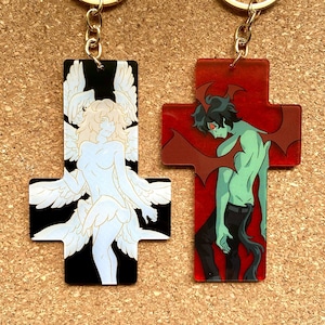 Può includere: Due portachiavi a forma di croce. Una croce è nera con una figura di angelo bianca con le ali. L'altra croce è rossa con una figura di diavolo verde con le ali.
