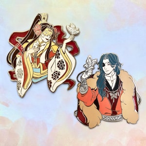 May include: Two enamel pins featuring anime-style characters. The left pin shows a figure in a cream and red robe with floral patterns and a mask. The right pin depicts a figure with blue hair, an eyepatch, and a tan fur-trimmed orange coat.