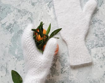 Può includere: Un paio di guanti bianchi lavorati a maglia, uno dei quali tiene un piccolo ramo di mandarini con foglie verdi. I guanti hanno un design strutturato e un aspetto soffice. Lo sfondo è grigio e bianco screziato.
