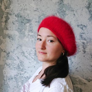 May include: A bright red beret made from a soft, fluffy material. The beret is worn by a person with dark hair and a white t-shirt. The background is a textured wall with shades of grey and blue.