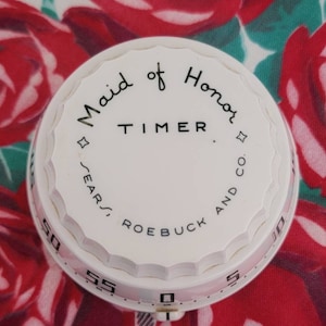 May include: A white, vintage kitchen timer with the words "Maid of Honor TIMER" in black script. The timer is from Sears, Roebuck and Co. and has a scalloped edge. The background is a floral pattern of red roses.
