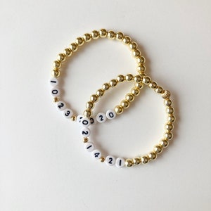 May include: Two gold beaded bracelets with white beads that have the numbers 2021 and 2023 printed on them.