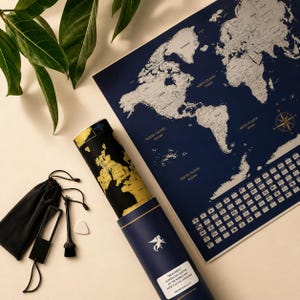 May include: A navy blue world map with silver detailing and a gold compass rose. A rolled-up map with gold foil continents is in a blue tube. Accessories include a black pouch, brush, and magnifying glass. The text on the tube reads "BEAUTIFUL ALTERNATIVE EDITION OF THE WORLD MAP WITH FLAGS & LANDMARKS".