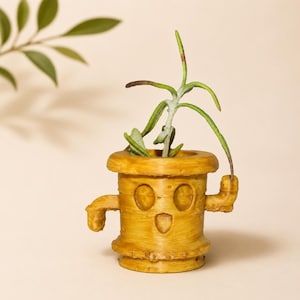 May include: A whimsical, yellow-brown planter shaped like a cartoon character, complete with arms, a smiling face, and a textured, wood-like finish. The planter holds a small green plant with long, slender leaves. The background is a soft, neutral color.