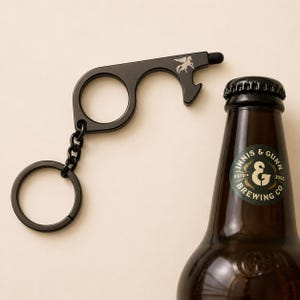 May include: A black multi-tool keychain with a bottle opener, stylus, and a pegasus design. The tool is next to a brown beer bottle with a green and white label.