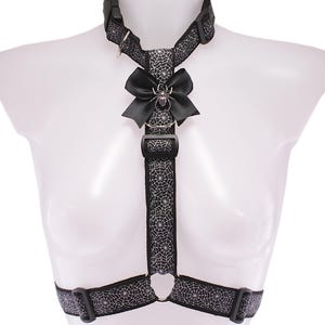 May include: Black harness with a spiderweb pattern and a black bow. The harness has adjustable straps and metal rings. A silver spider charm is centered on the bow. The harness is displayed on a white mannequin.