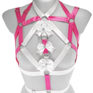 Op de afbeelding: Een roze en wit harnas met strikaccenten. Het harnas heeft roze bandjes met goudkleurige metalen hardware en witte bandjes. Witte strikken met strass-middens zijn aan het harnas bevestigd.