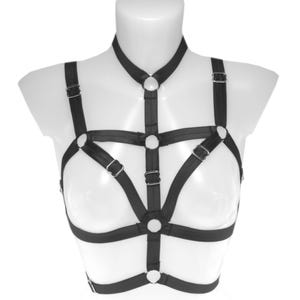 May include: Black body harness with adjustable straps and metal hardware. The harness features a halter neck, shoulder straps, and a chest design. The design includes multiple O-rings and adjustable buckles for a secure fit. The harness is displayed on a white mannequin.