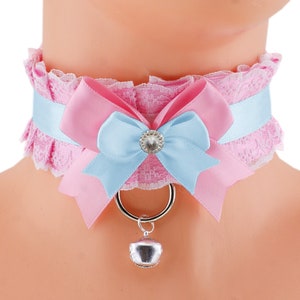 Könnte beinhalten: Rosa Spitzenhalsband mit hellblauem Satinband und rosa Satinschleife. Die Schleife ist mit einem Strassstein verziert. Ein silberner Ring und eine Glocke hängen an dem Halsband.