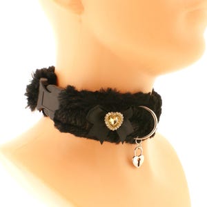 Op de afbeelding: Zwarte nep bont choker met een zwart satijnen lint en een hartvormige hanger met een heldere strass. De choker heeft een zilveren metalen ring en een zilveren hangslot hanger.