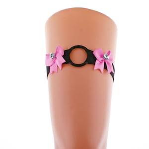 May include: Black elastic garter with a central ring design, adorned with two pink satin bows. Each bow features a sparkling rhinestone. The garter is displayed on a mannequin leg against a white background.