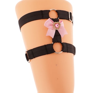 May include: Black garter with two bands and silver-tone metal rings. A pink bow with a heart-shaped jewel is centered on the garter. The garter is displayed on a mannequin leg against a white background.