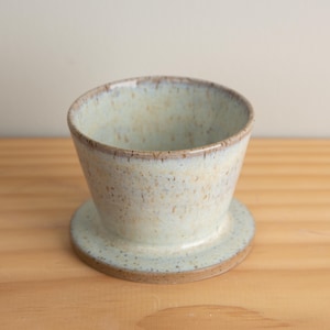 May include: A ceramic pour-over coffee dripper in a speckled, light blue glaze. The dripper has a conical shape with a wide base and a small, circular platform. The rim and base have a darker, brown color. The dripper sits on a wooden surface.
