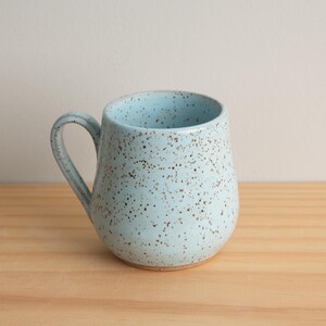 May include: A light blue ceramic mug with a speckled brown glaze. The mug has a rounded body and a handle.