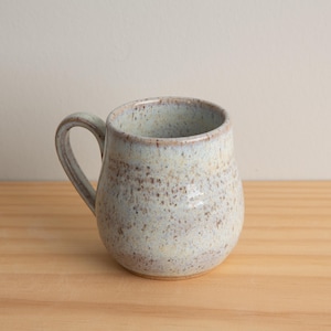 Puede incluir: Una taza de cerámica blanca con un esmalte moteado y un cuerpo redondeado. La taza tiene un asa y es perfecta para disfrutar de una bebida caliente.