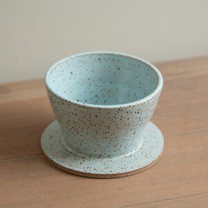 Può includere: Una caffettiera a versamento in ceramica blu chiaro macchiettata con un piattino abbinato. La caffettiera ha una forma a ciotola ampia e aperta e poggia su una base circolare. La ceramica presenta un motivo macchiettato.