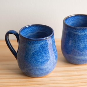 May include: Two blue ceramic mugs with a speckled glaze. The mugs have a rounded shape and a handle.