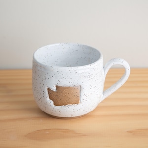Può includere: Tazza in ceramica bianca maculata con un ritaglio marrone del contorno dello stato di Washington. La tazza ha una forma arrotondata con un manico e si staglia su uno sfondo chiaro.