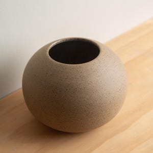 May include: A spherical, speckled ceramic vase in a neutral tan color. The vase has a wide opening and a dark interior. The surface has a textured appearance. This decorative vase is suitable for home decor.