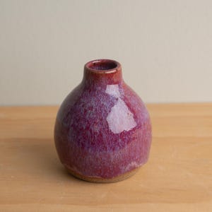 May include: A small, round ceramic vase with a narrow neck. The vase has a glossy finish with a blend of red and purple hues. The base of the vase is a lighter, natural clay colour. The vase is on a wooden surface.