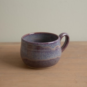 May include: A handcrafted ceramic mug with a rounded form and a handle. The mug has a speckled, layered design in shades of purple and blue. The interior is a darker purple, and it rests on a wooden surface.