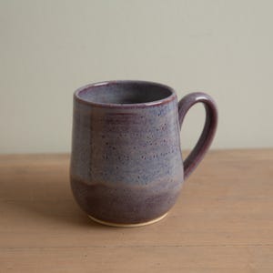 May include: A handmade ceramic mug with a rounded body and a sturdy handle. The mug features a speckled, two-tone glaze in shades of purple and blue, with a lighter base. The mug is sitting on a wooden surface.