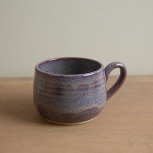 May include: A handmade ceramic mug with a rounded body and a handle. The mug features a blend of purple and blue hues with a speckled texture. The interior and handle are a darker shade of purple. The mug sits on a light-colored base.