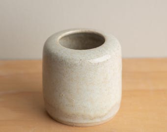 May include: A small, off-white ceramic vase with a cylindrical shape and a rounded top. The vase has a smooth, speckled texture and a small opening at the top. The vase is sitting on a wooden surface.